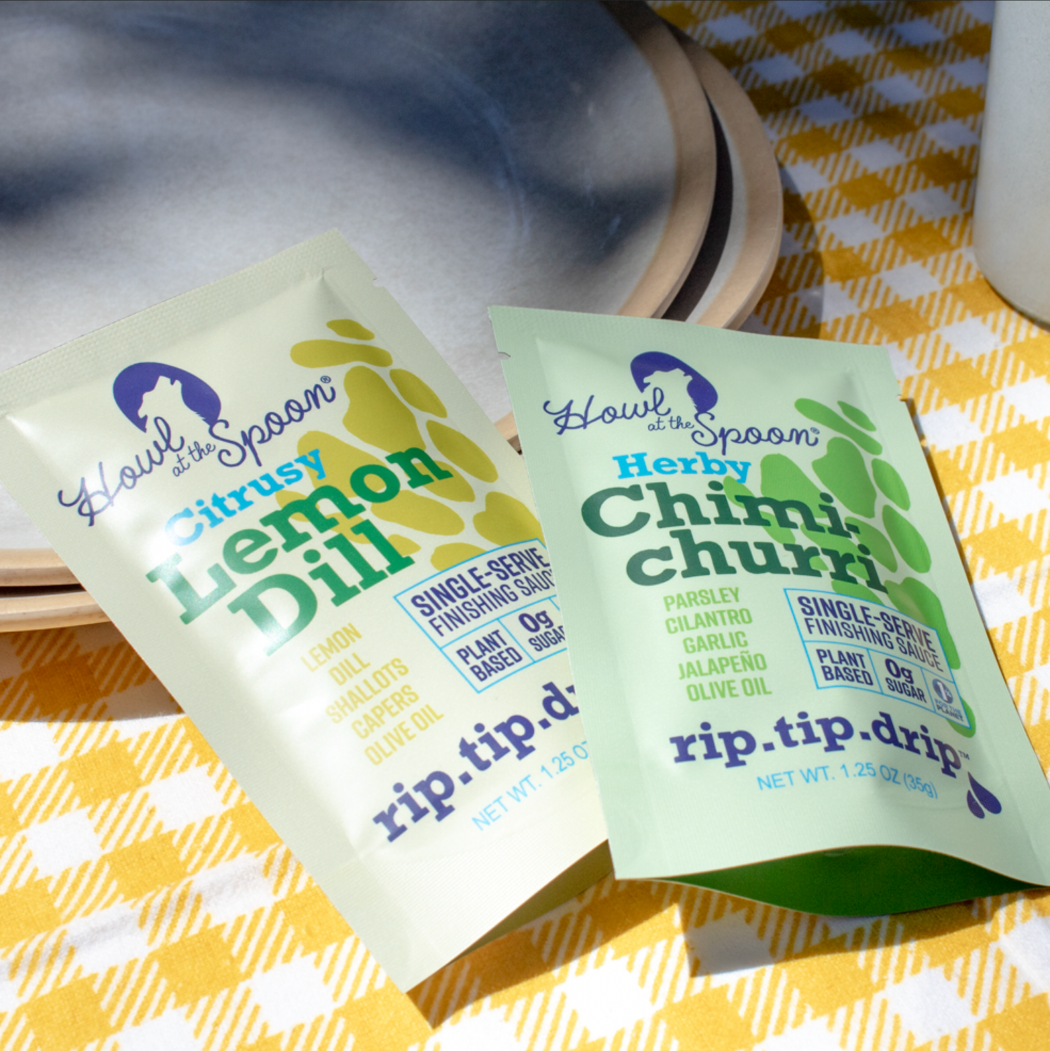 Howl at the Spoon’s Lemon Dill and Chimichurri single-serve sauces at an outdoor picnic setting, placed beside fresh veggies and snacks on a blanket.